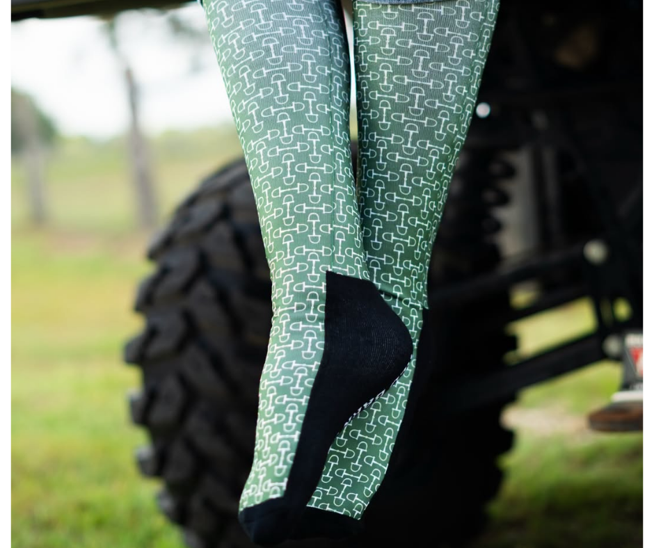 Green and white red barn socks worn by a person sitting on a vehicle outdoors, with a large black tire and blurred grass in the background.
