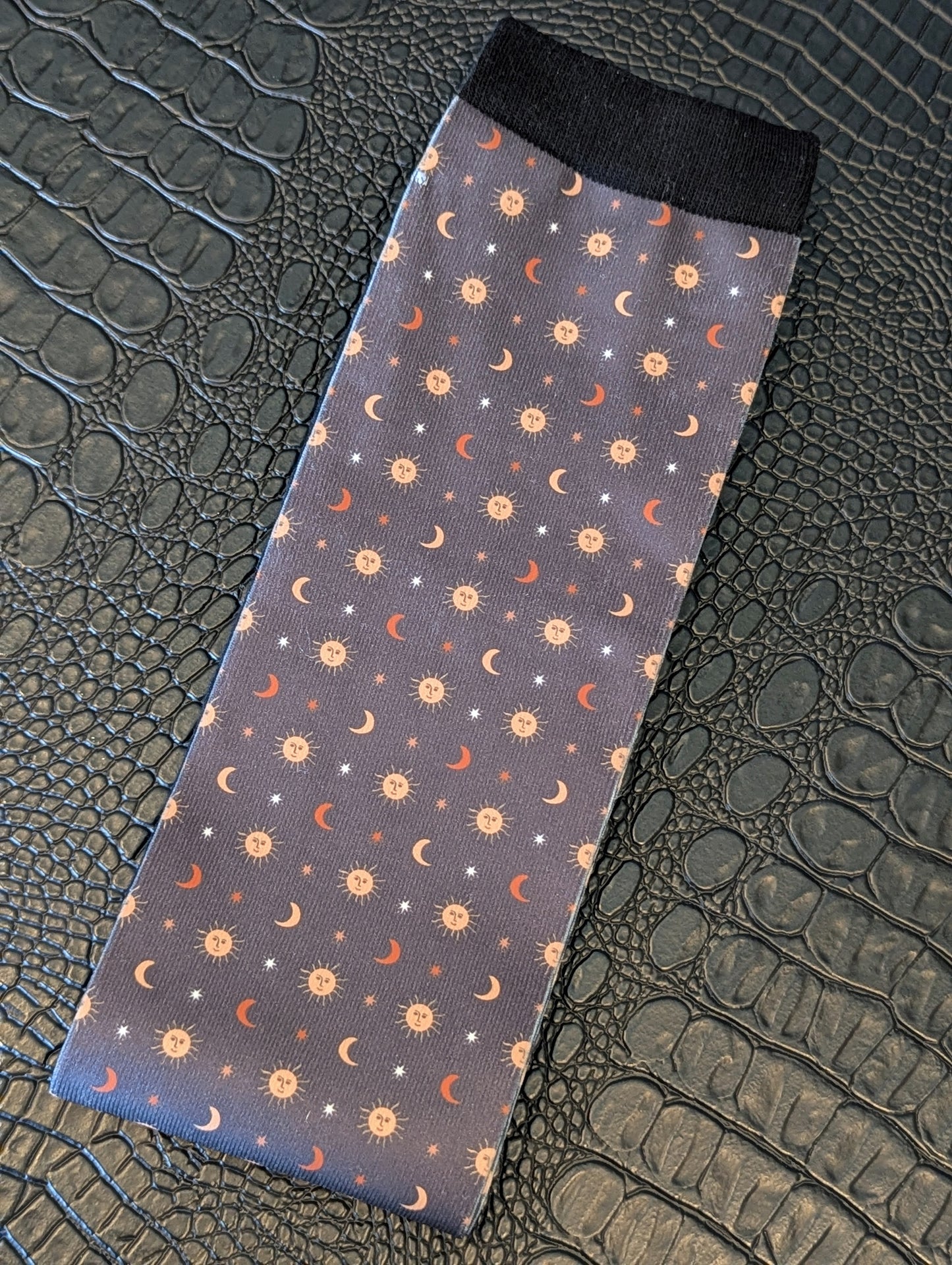 Navy sock with celestial sun, moon, and star pattern and black cuff displayed on a textured black surface