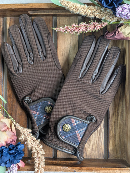   
Brown tobacco riding gloves with leather palms and breathable mesh backs, displayed on a wooden tray with a Field Plaid wrist tab and surrounded by dried florals.