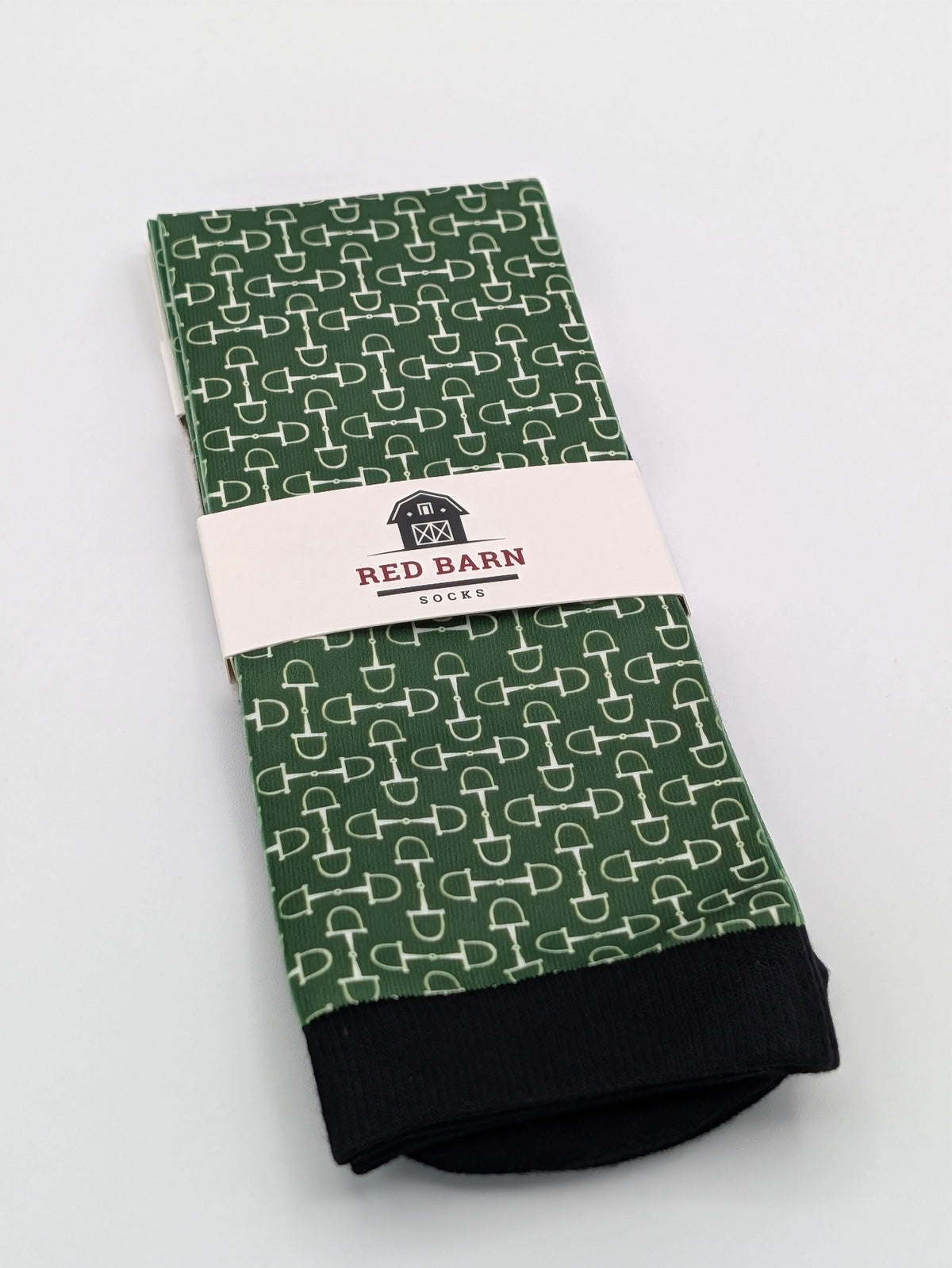 A pair of folded green with white bit patterned socks with black cuffs, wrapped in a white paper band labeled 'RED BARN SOCKS', on a light gray background.