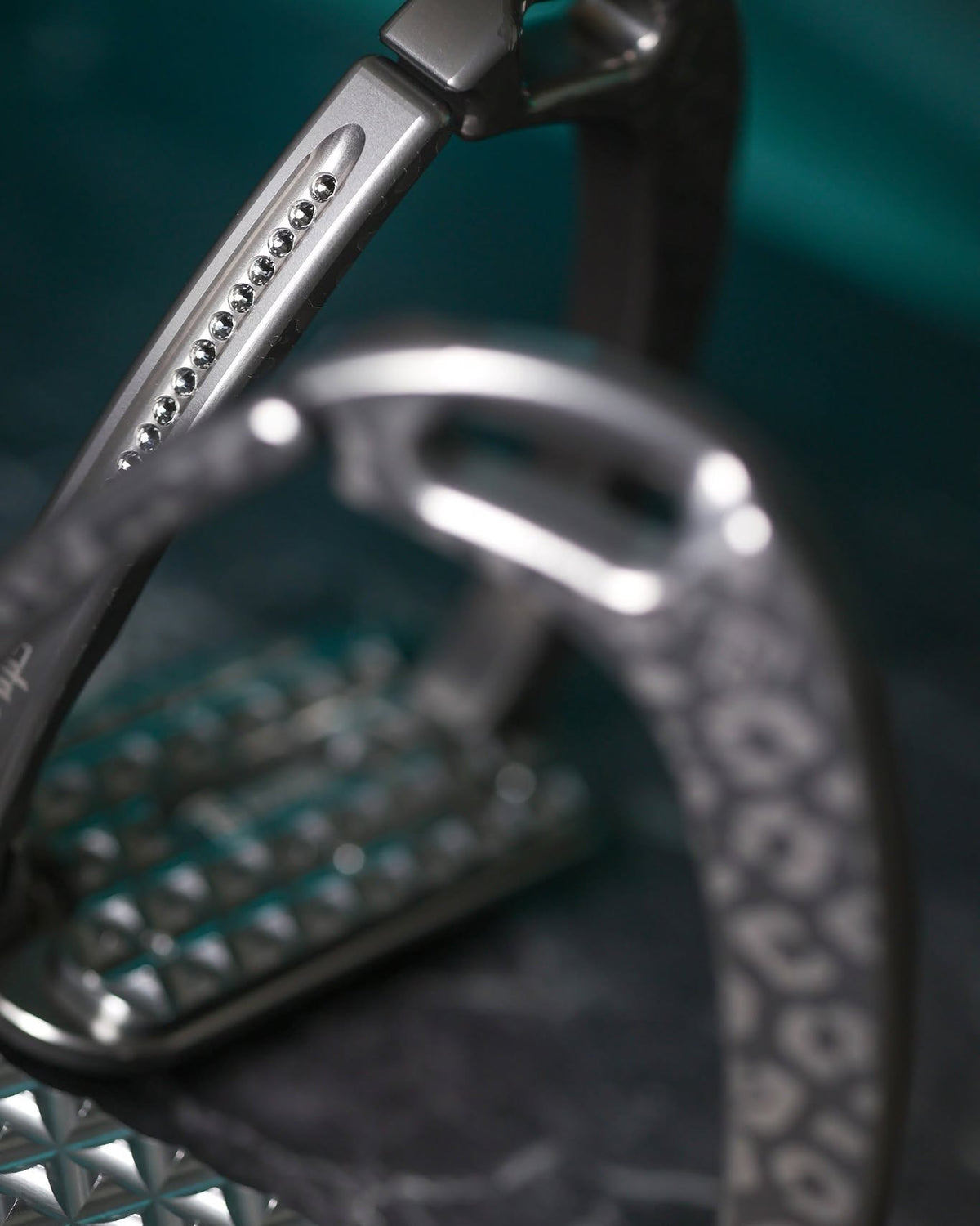 Titanium Italian‑made safety stirrups with a matte leopard pattern and dark silver Swarovski crystals, shown against a teal background with a close‑up of the textured tread.