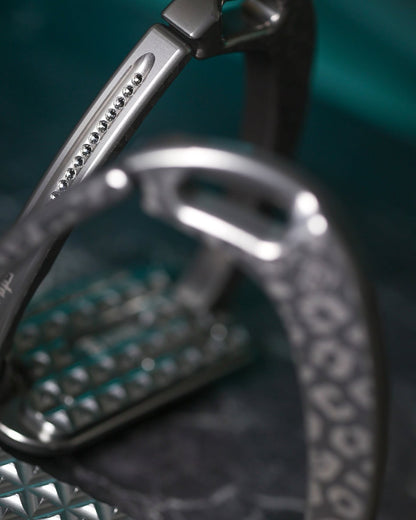 Titanium Italian‑made safety stirrups with a matte leopard pattern and dark silver Swarovski crystals, shown against a teal background with a close‑up of the textured tread.