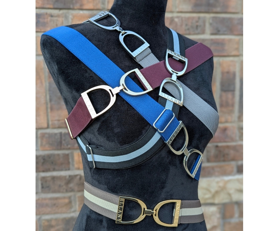 A black mannequin torso layered with multiple wide Ellany elastic belts in fall colors—petrol blue, burgundy, gray, and taupe—each featuring a polished silver stirrup buckle, displayed against a brick wall.
