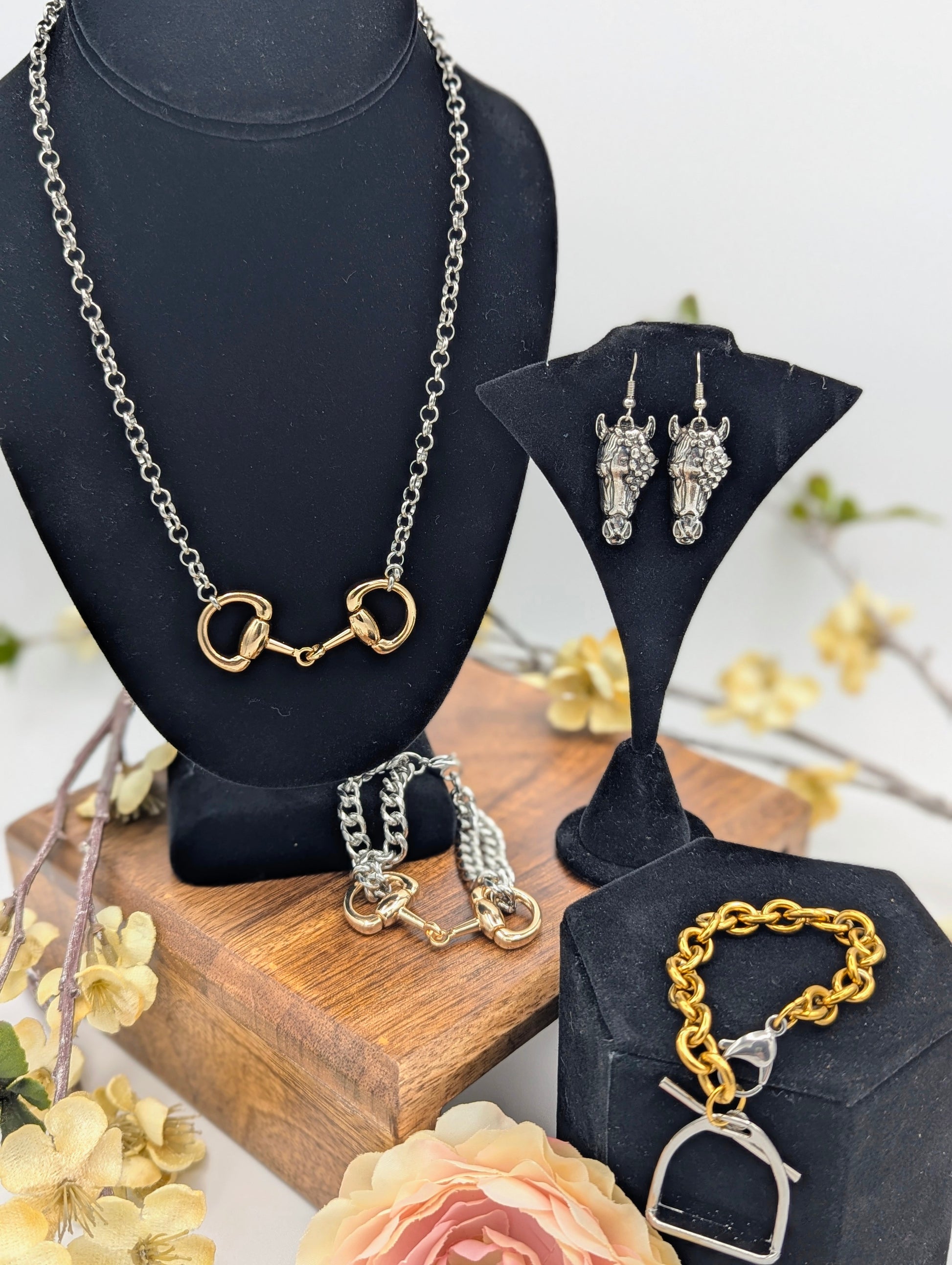 Equestrian jewelry collection featuring a two‑tone snaffle bit necklace, horse head dangle earrings, chunky snaffle bit bracelet, and two‑tone stirrup bracelet displayed on black stands with floral accents.