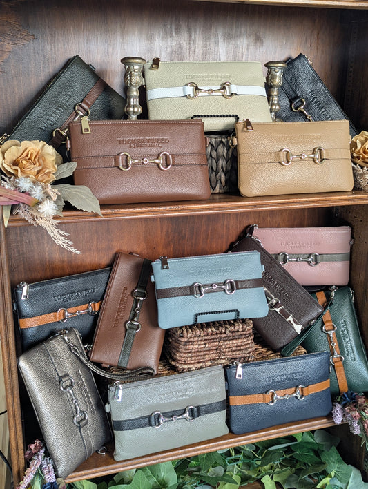 A collection of Tucker Tweed horsebit-embellished zip-top leather wallets and clutch bags in various colors including brown, beige, pink, blue, gray, and navy, displayed on wooden shelves with decorative dried flowers.