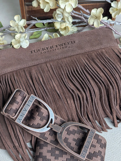 A brown suede fringe pouch with a matching patterned belt and buckle, displayed on a light surface with decorative faux flowers in the background. The pouch is embossed with 'TUCKER TWEED EQUESTRIAN' and the buckle is engraved with 'ELLANY'.