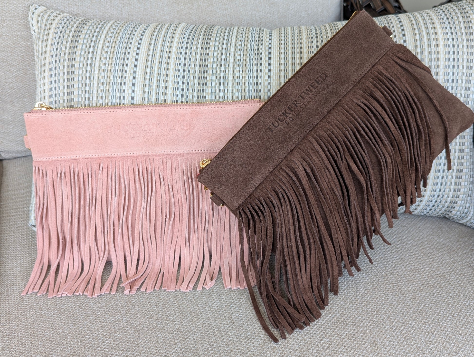Two fringe pouches or clutch-style bags displayed on a textured sofa with a striped pillow. One is light blush pink suede with long fringe and a small gold zipper pull; the other is medium brown suede with long fringe and a gold zipper pull.