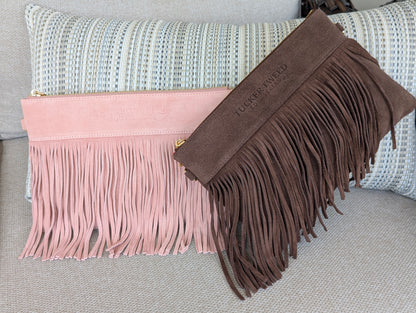 Two fringe pouches or clutch-style bags displayed on a textured sofa with a striped pillow. One is light blush pink suede with long fringe and a small gold zipper pull; the other is medium brown suede with long fringe and a gold zipper pull.
