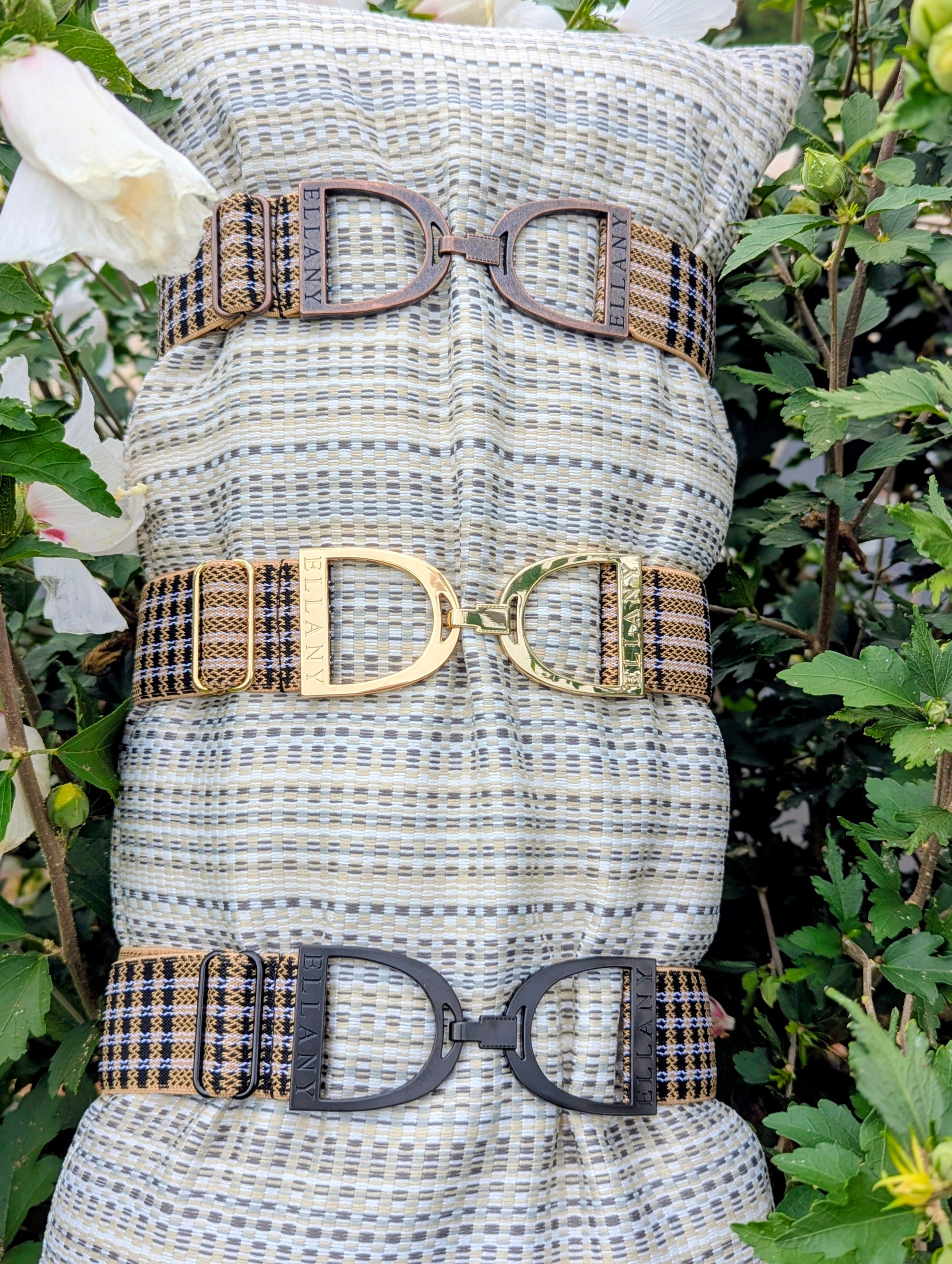 Three Limited Edition 5/A Baker × Ellany elastic belts displayed on a striped cushion among greenery and white flowers, each featuring signature Baker plaid and a different metal buckle finish—copper, yellow‑gold, and matte black—with “ELLANY” engraved on each buckle.