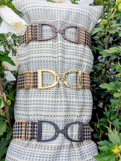 Three Limited Edition 5/A Baker × Ellany elastic belts displayed on a striped cushion among greenery and white flowers, each featuring signature Baker plaid and a different metal buckle finish—copper, yellow‑gold, and matte black—with “ELLANY” engraved on each buckle.