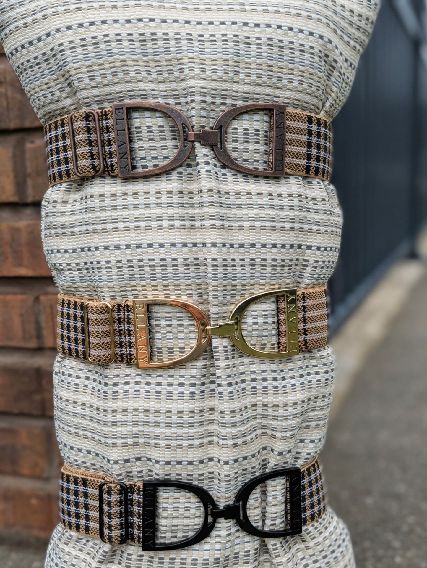 Three Limited Edition 5/A Baker × Ellany elastic belts displayed on a woven mannequin stand, each featuring signature Baker plaid and a different metal buckle finish—copper, gold, and matte black—with “ELLANY” engraved on each buckle.