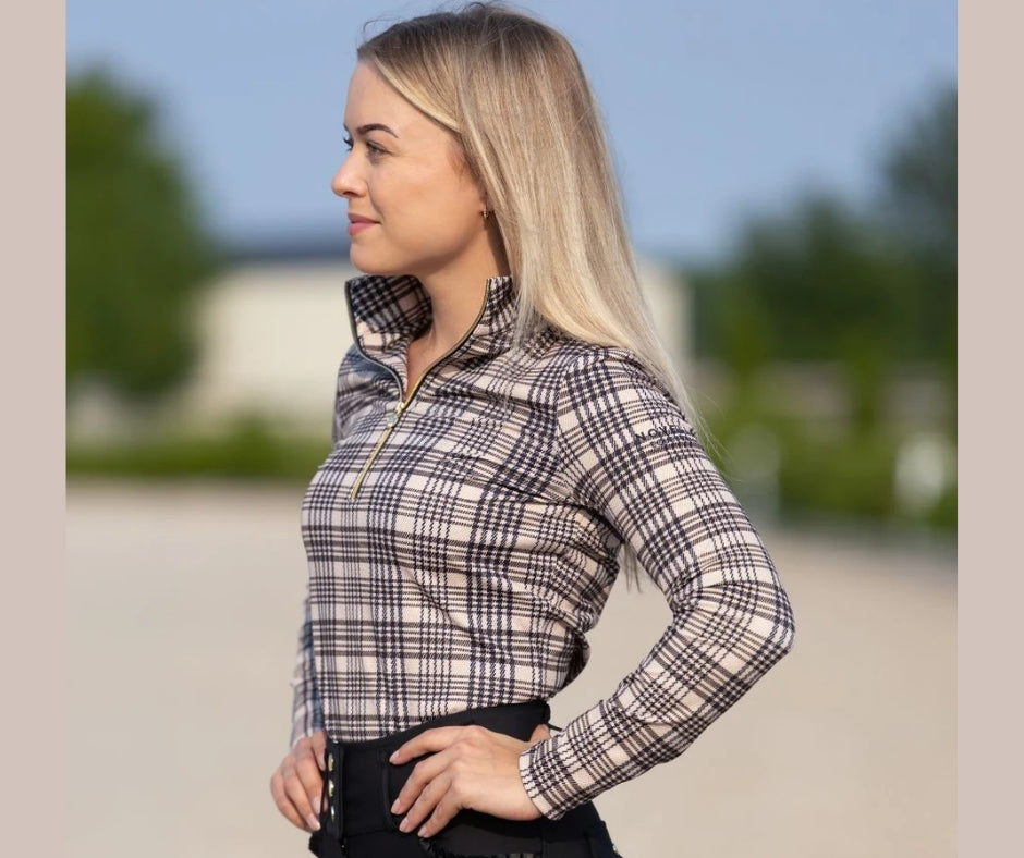 Woman wearing the 5/A Baker × Novella plaid mid‑weight quarter zip, shown from the side in an outdoor equestrian setting.