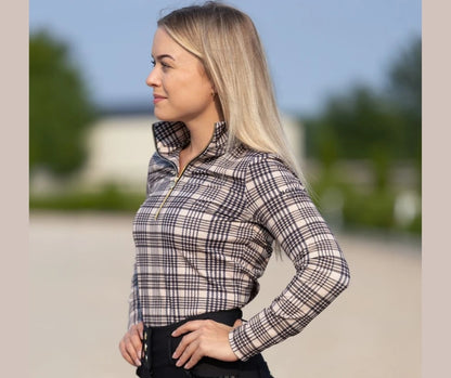 Woman wearing the 5/A Baker × Novella plaid mid‑weight quarter zip, shown from the side in an outdoor equestrian setting.