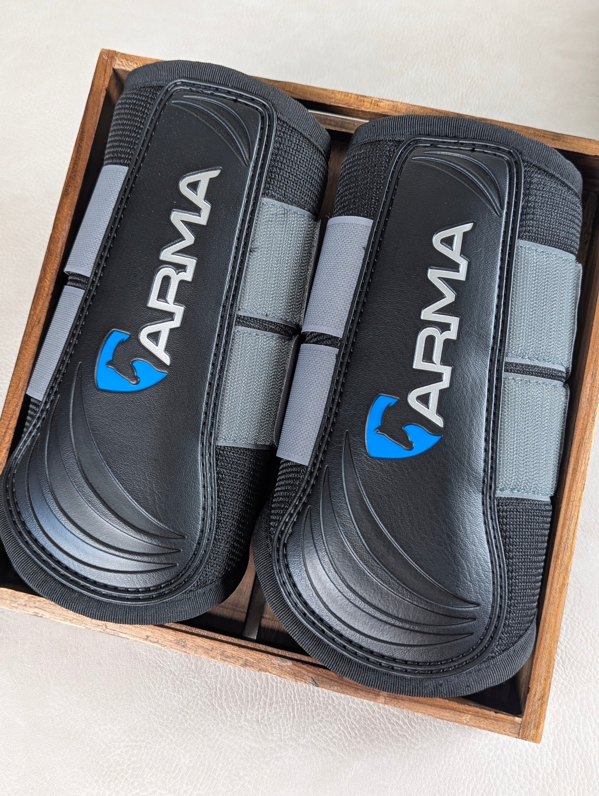 A pair of black and gray ARMA brushing boots displayed inside a wooden box, showing the contoured protective shell and branded detailing.