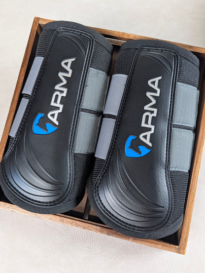 A pair of black and gray ARMA brushing boots displayed inside a wooden box, showing the contoured protective shell and branded detailing.