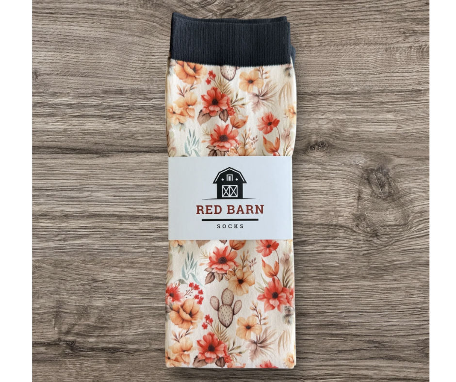 A pair of folded socks with a floral print and a black ribbed cuff, wrapped in a white paper band labeled 'RED BARN SOCKS', displayed on a gray wood background.