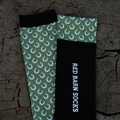 Sage green horseshoe‑pattern sock paired with a black Red Barn logo sock laid out on a tree stump