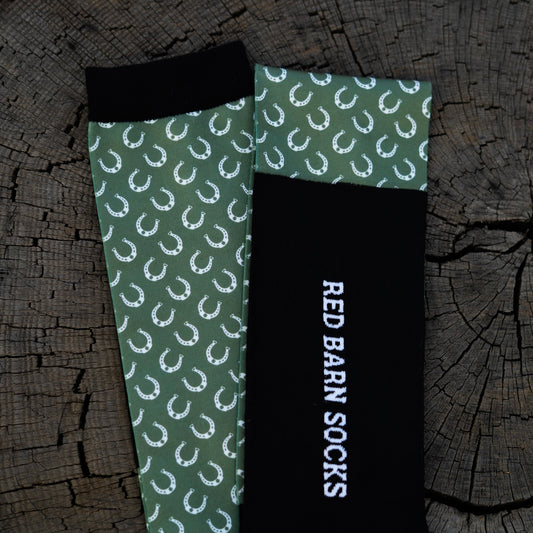 Sage green horseshoe‑pattern sock paired with a black Red Barn logo sock laid out on a tree stump