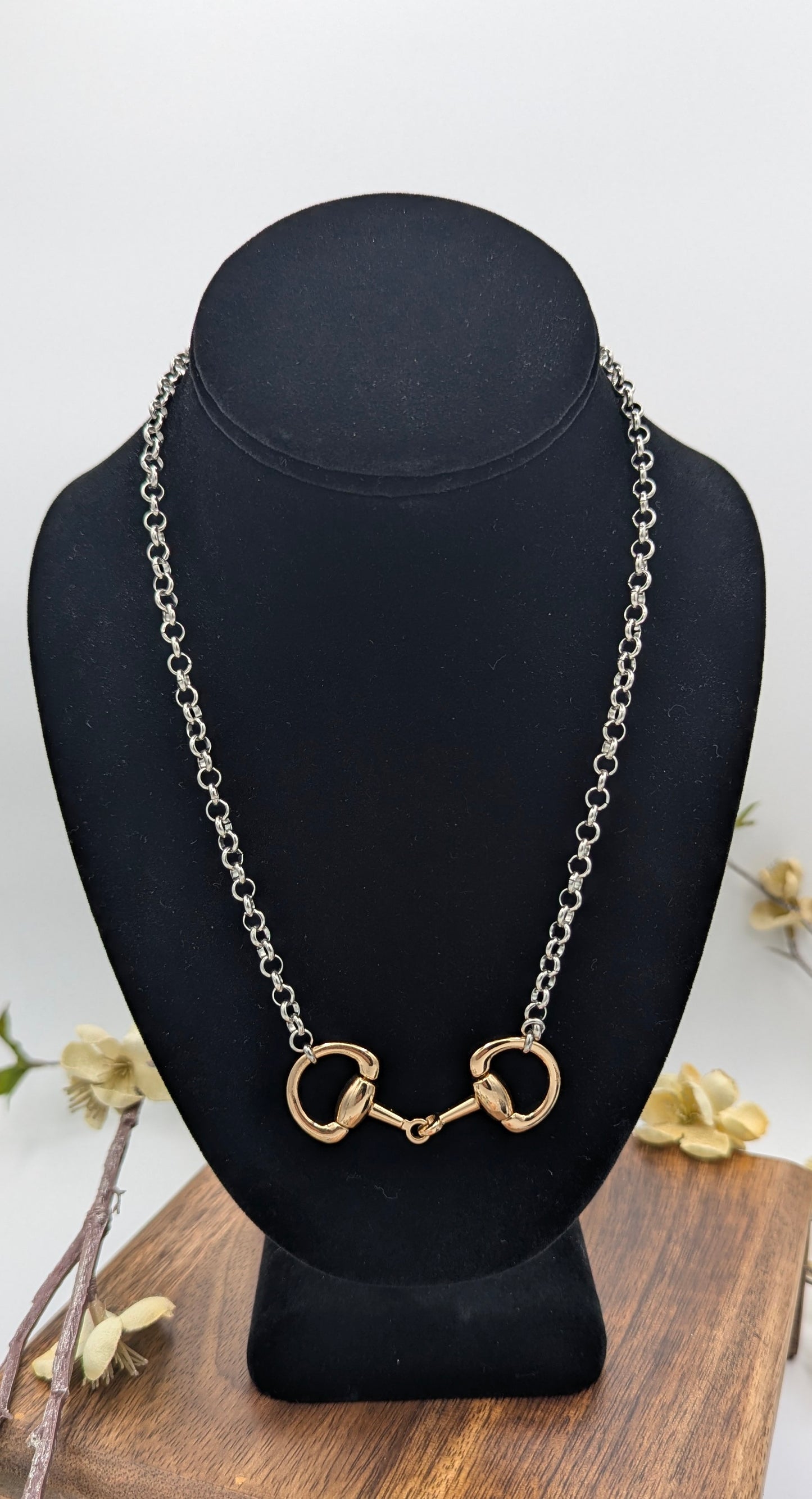   
Silver‑tone chain necklace with a gold‑tone snaffle bit centerpiece displayed on a black velvet jewelry bust, accented by small cream flowers on a wooden surface.