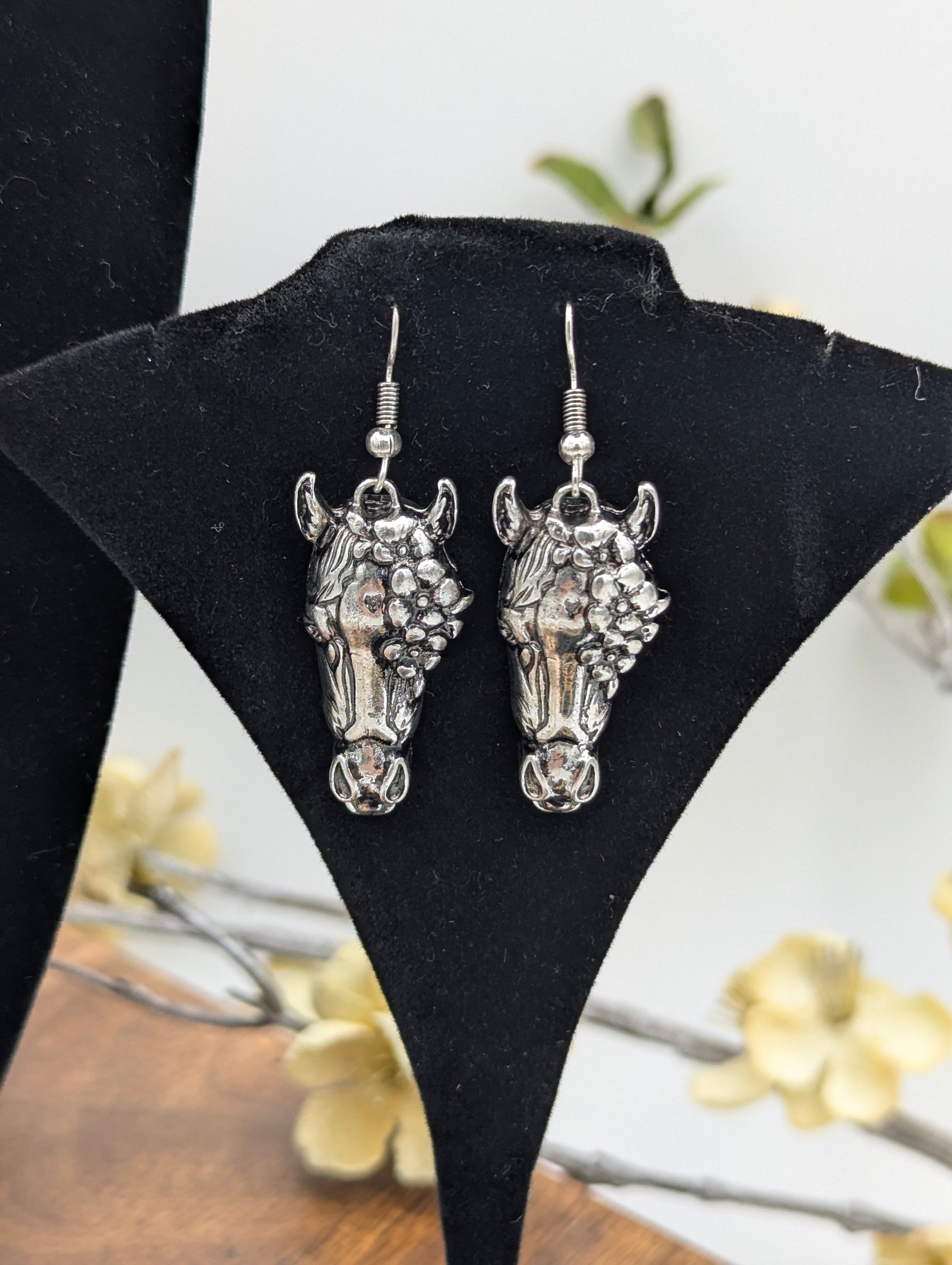 Silver‑tone dangle earrings shaped as detailed horse heads with floral accents, displayed on a black velvet stand with soft floral décor in the background.