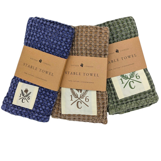 Three textured cotton dish towels in blue, brown, and green, each wrapped with a kraft paper band labeled 'STABLE TOWEL' and a small sewn patch with a monogram logo, shown on a white background.