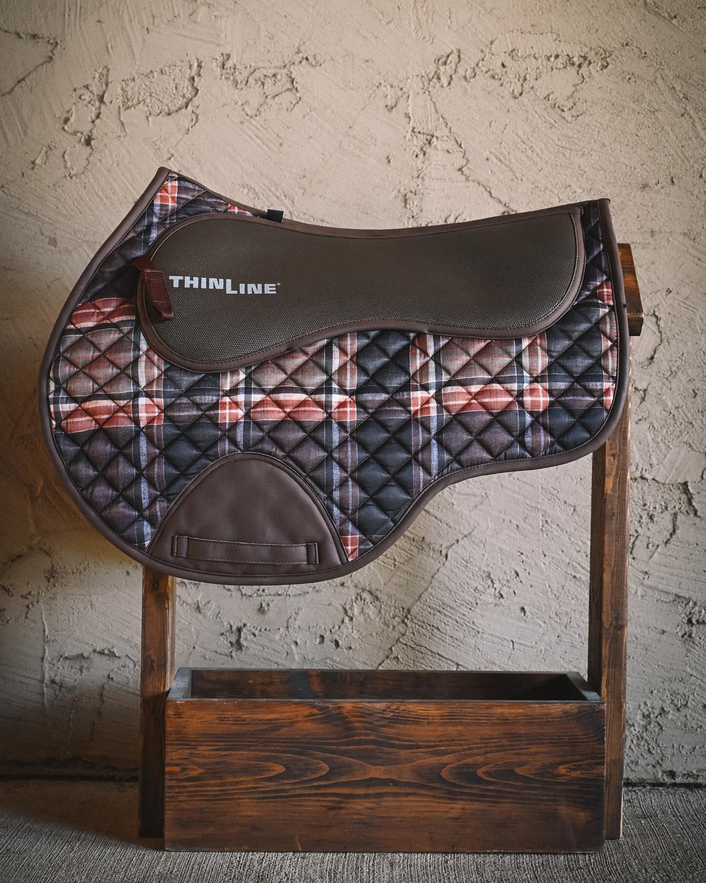 Field Plaid close contact saddle pad displayed on a wooden stand, featuring a quilted plaid body, smooth brown top panel with ThinLine branding, and cognac leatherette binding.