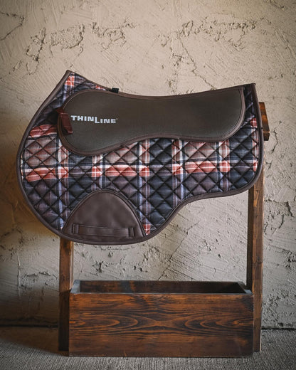 Field Plaid close contact saddle pad displayed on a wooden stand, featuring a quilted plaid body, smooth brown top panel with ThinLine branding, and cognac leatherette binding.