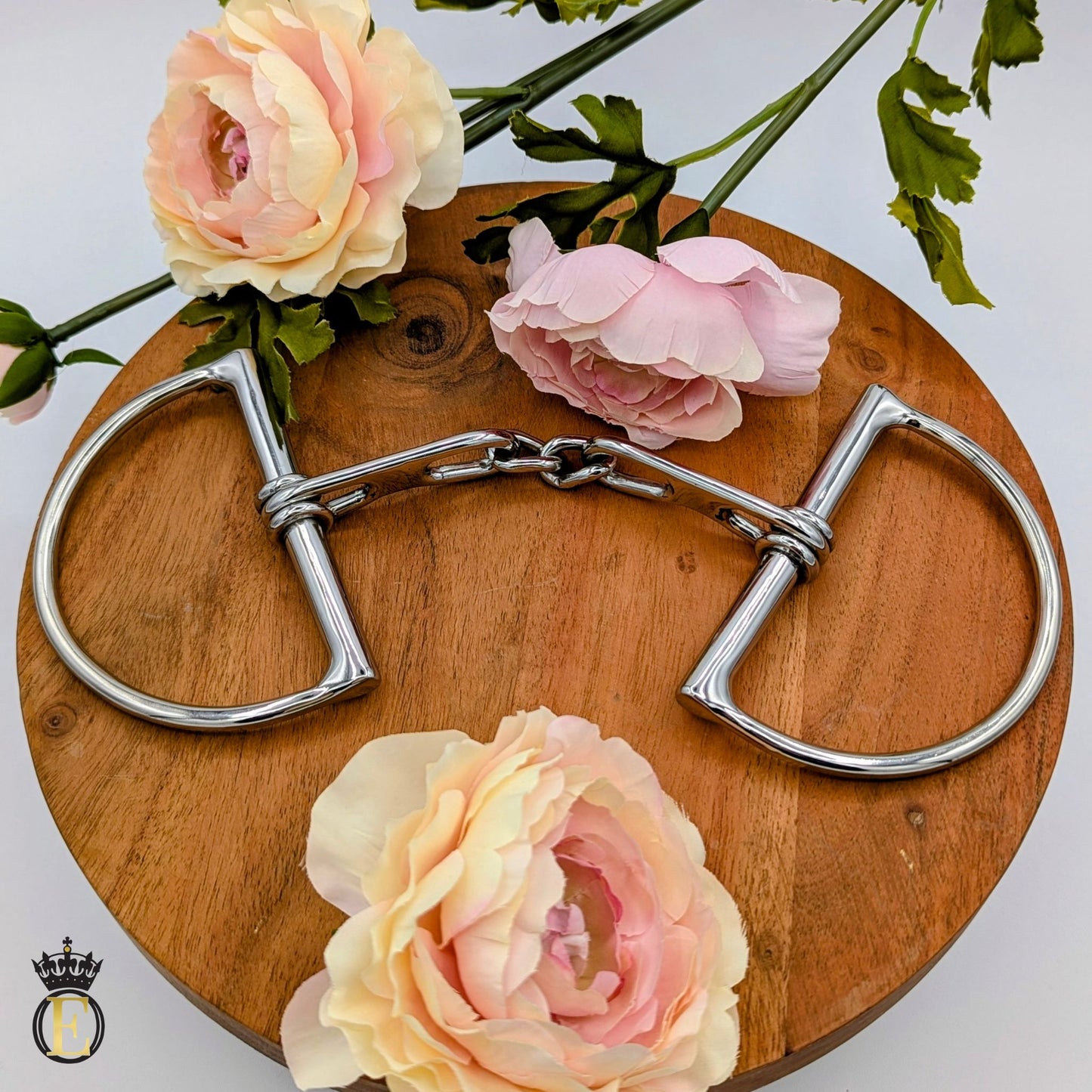 A polished stainless peter pletcher teardrop dee horse bit with a curved double-ring design and a short shank, displayed on a round wooden board with pink and cream roses.