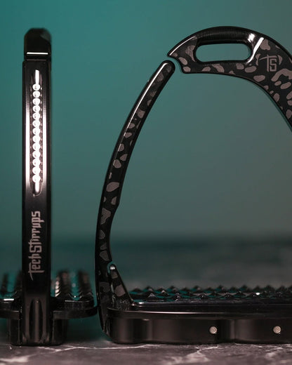 Black limited‑edition Italian safety stirrups with leopard finish and clear Swarovski crystals, shown on a marble surface with a teal gradient backdrop.