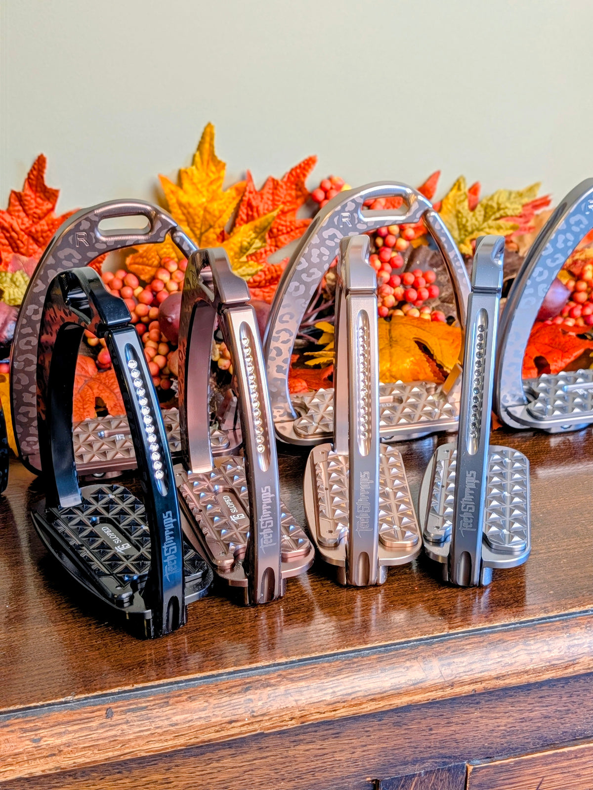 Four pairs of Tech Stirrups Venice Sloped EVO in assorted limited‑edition leopard finishes arranged on a wooden surface with fall leaves in the background; textured tread and Swarovski‑accented frames visible.