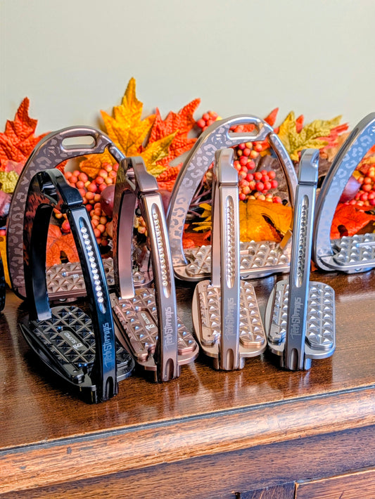 Four pairs of Tech Stirrups Venice Sloped EVO in assorted limited‑edition leopard finishes arranged on a wooden surface with fall leaves in the background; textured tread and Swarovski‑accented frames visible.