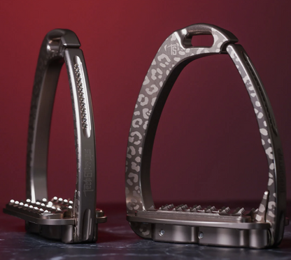 Rose gold Italian‑made safety stirrups with a soft leopard pattern and powder‑shade Swarovski crystals, shown against a red gradient background.