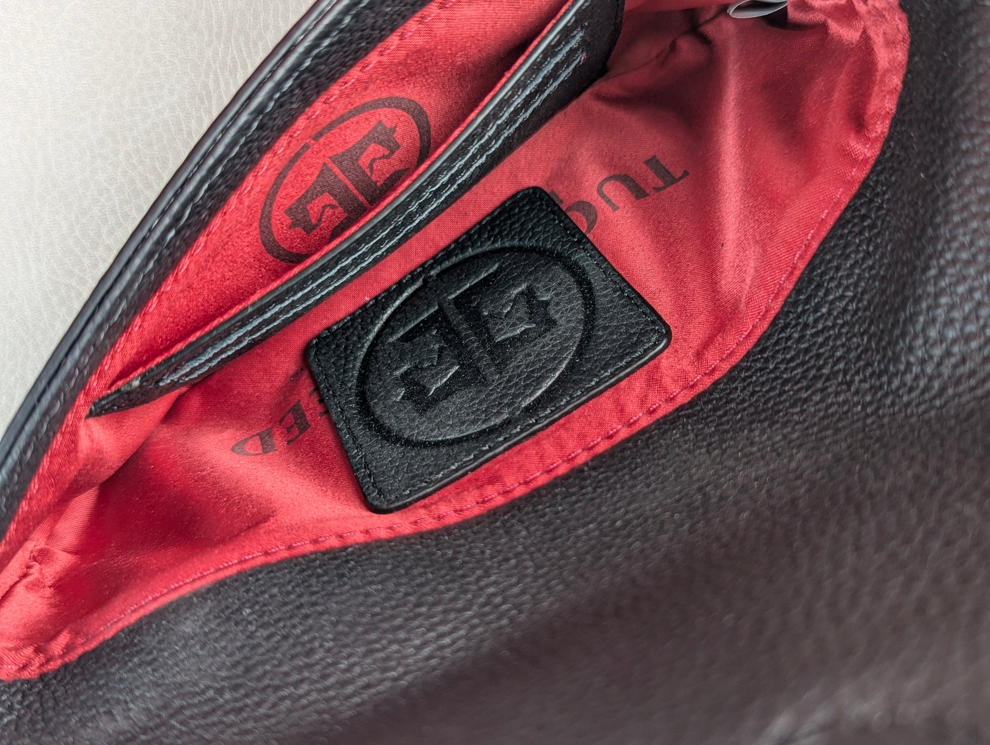 Close-up of a black textured leather tucker tweed stablemate belt bag interior with a bright red satin lining and a black leather patch featuring an embossed monogram logo.