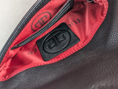 Close-up of a black textured leather tucker tweed stablemate belt bag interior with a bright red satin lining and a black leather patch featuring an embossed monogram logo.