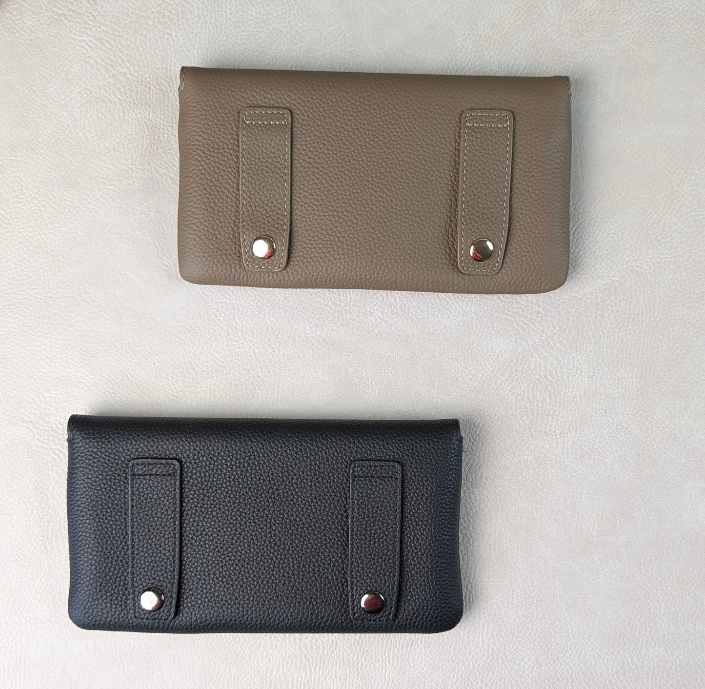 Two pebbled leather tucker tweed stablemate belt bag accessories laid flat on a light textured background, one in taupe and one in black, each with two vertical strap details and metal snap hardware for belt loops.