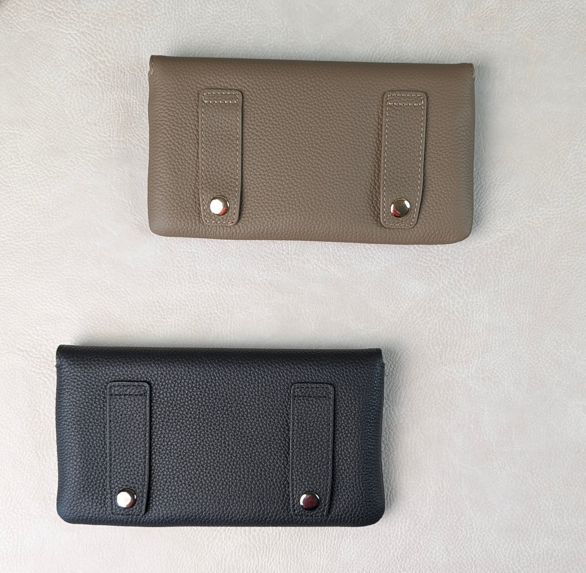 Two pebbled leather tucker tweed stablemate belt bag accessories laid flat on a light textured background, one in taupe and one in black, each with two vertical strap details and metal snap hardware for belt loops.