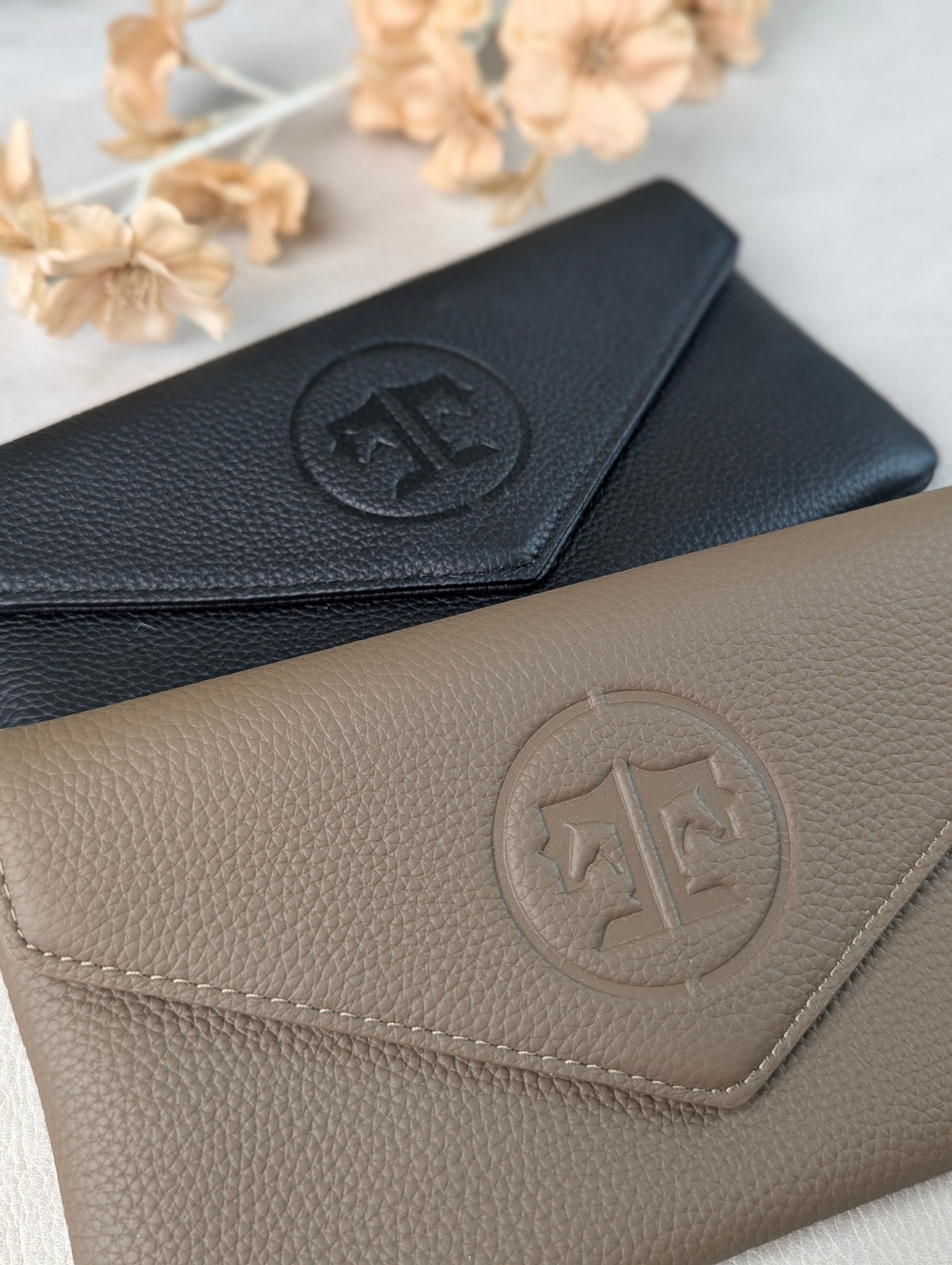 Two pebbled leather envelope-style belt bags  in black and stone, arranged diagonally on a light surface with dried flowers in the background. Each piece has an embossed tucker tweed circular monogram logo on the flap.
