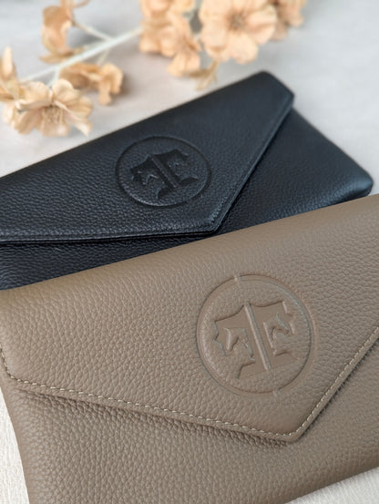Two pebbled leather envelope-style belt bags  in black and stone, arranged diagonally on a light surface with dried flowers in the background. Each piece has an embossed tucker tweed circular monogram logo on the flap.