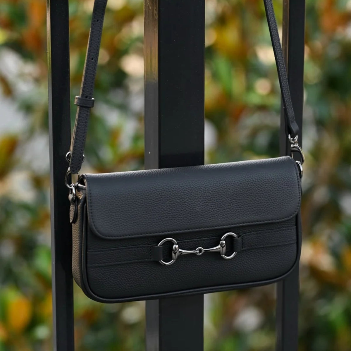 Black pebbled‑leather equestrian shoulder bag hanging on a railing, featuring a structured flap design with silver horsebit hardware and an adjustable leather strap, shown in an outdoor setting with softly blurred foliage.