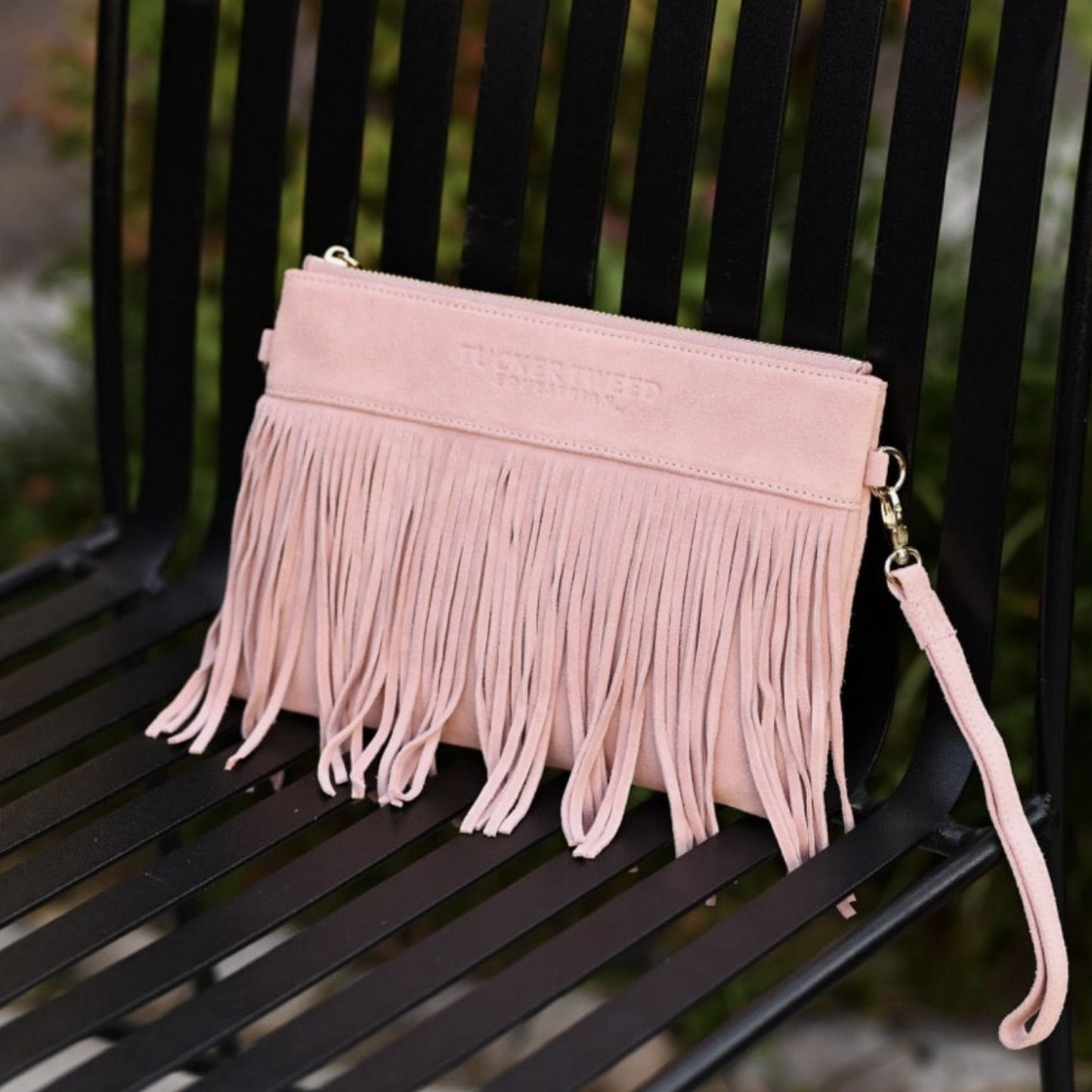 A light pink suede-leather clutch bag with long fringe and a wrist strap, resting on a black metal bench.