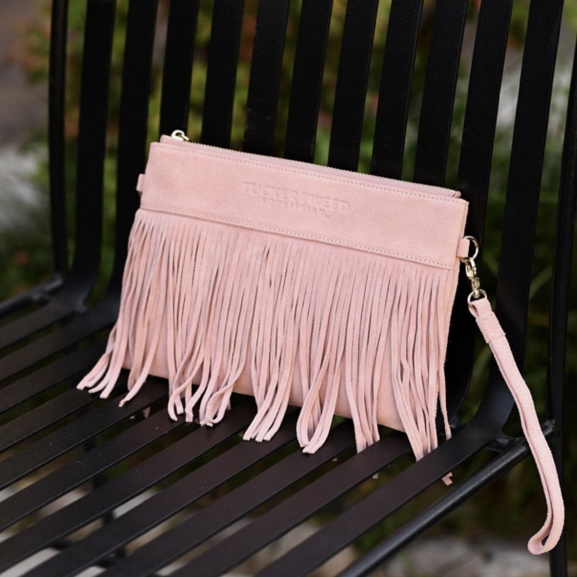 A light pink suede-leather clutch bag with long fringe and a wrist strap, resting on a black metal bench.