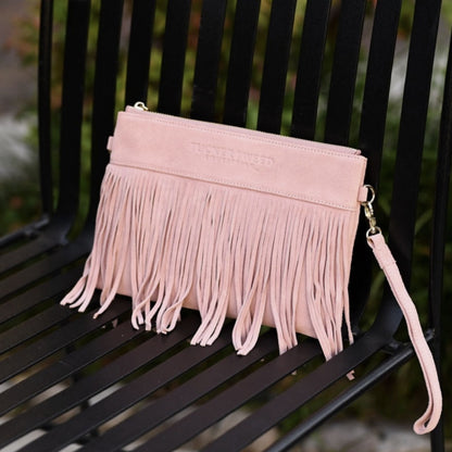 A light pink suede-leather clutch bag with long fringe and a wrist strap, resting on a black metal bench.