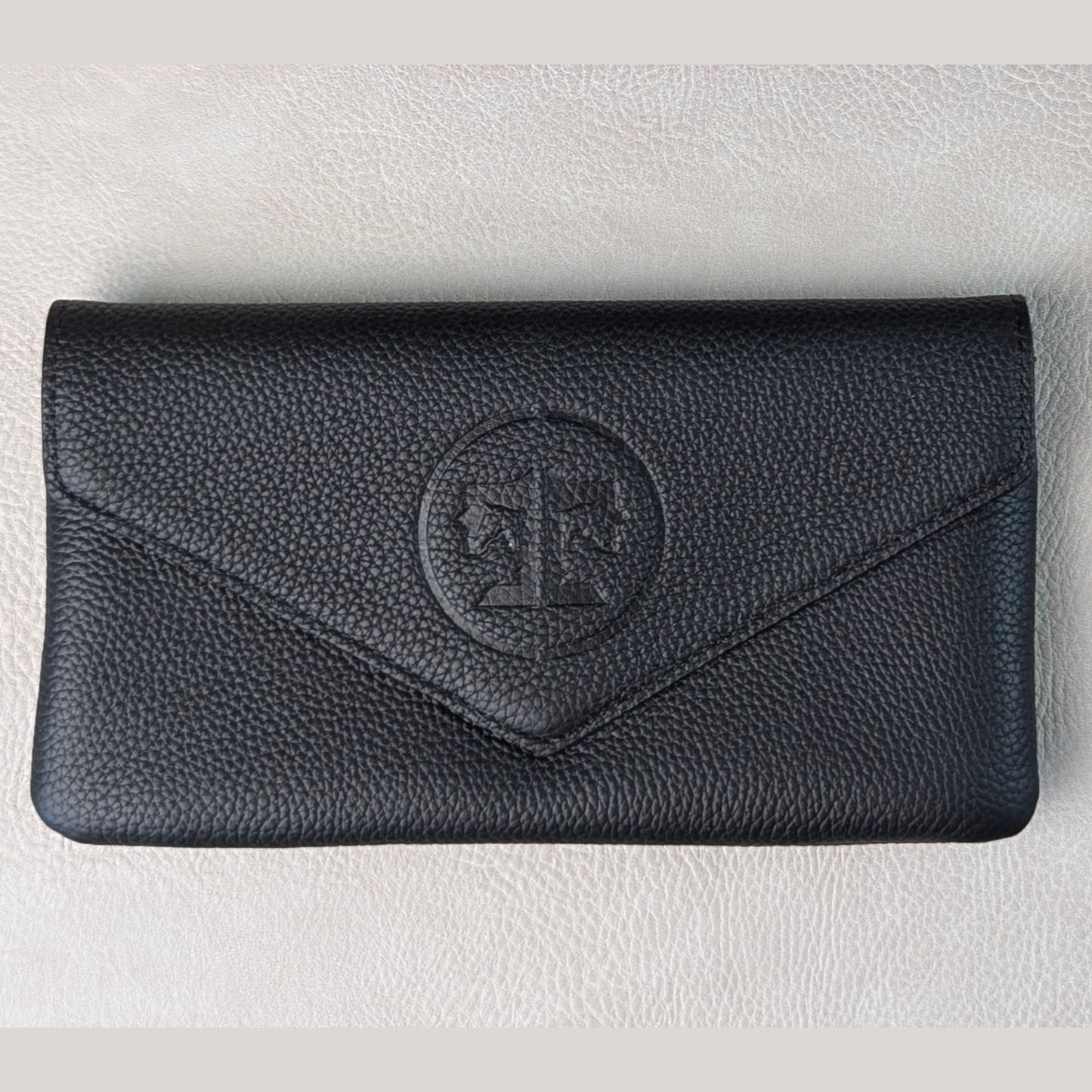 A black pebbled leather envelope-style wallet with a circular embossed monogram logo on the front, displayed on a light textured surface.