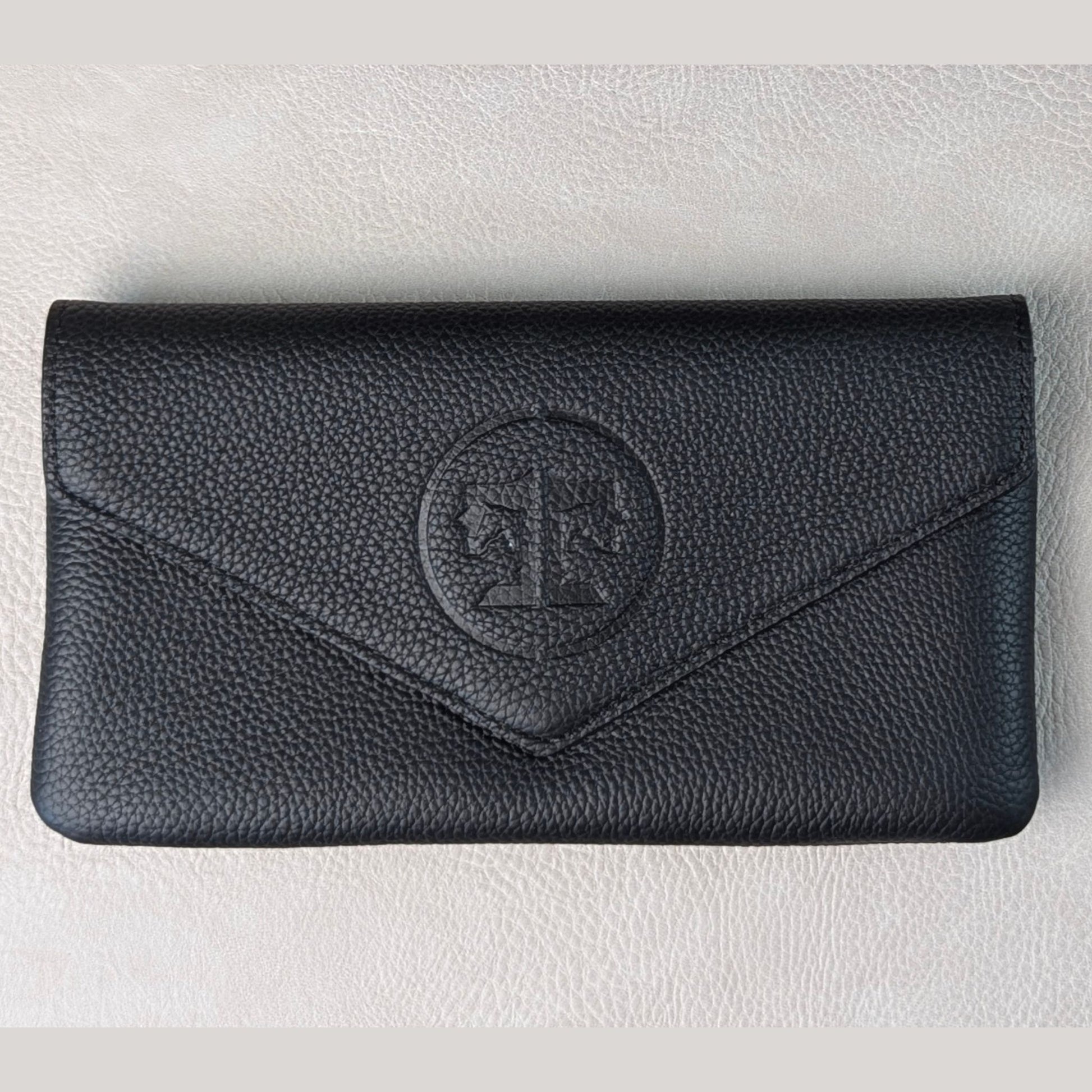 A black pebbled leather envelope-style wallet with a circular embossed monogram logo on the front, displayed on a light textured surface.