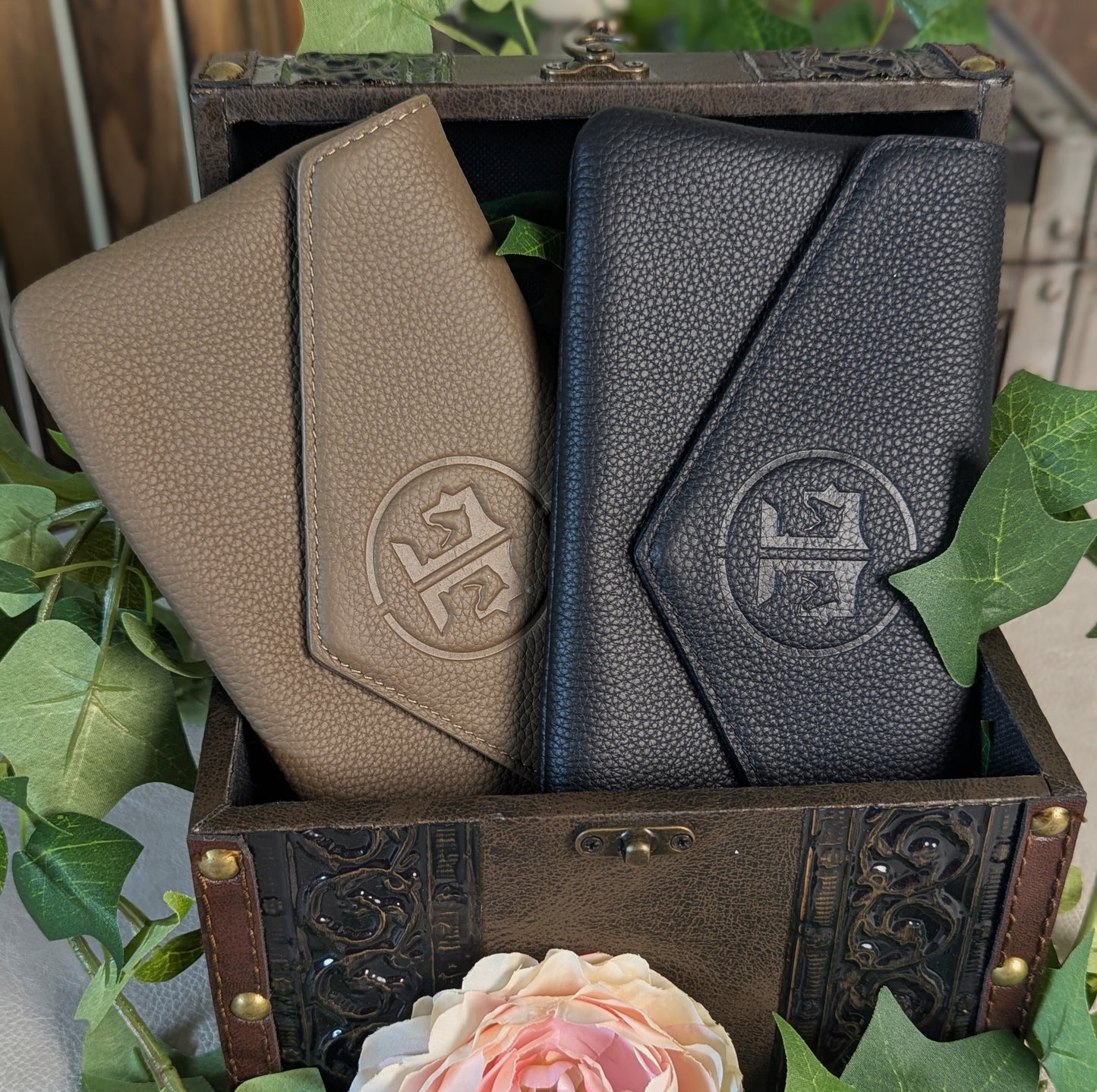 Two textured leather wallets with an embossed monogram logo, one in taupe and one in black, displayed inside an ornate vintage-style wooden chest surrounded by green leaves and a pink rose.