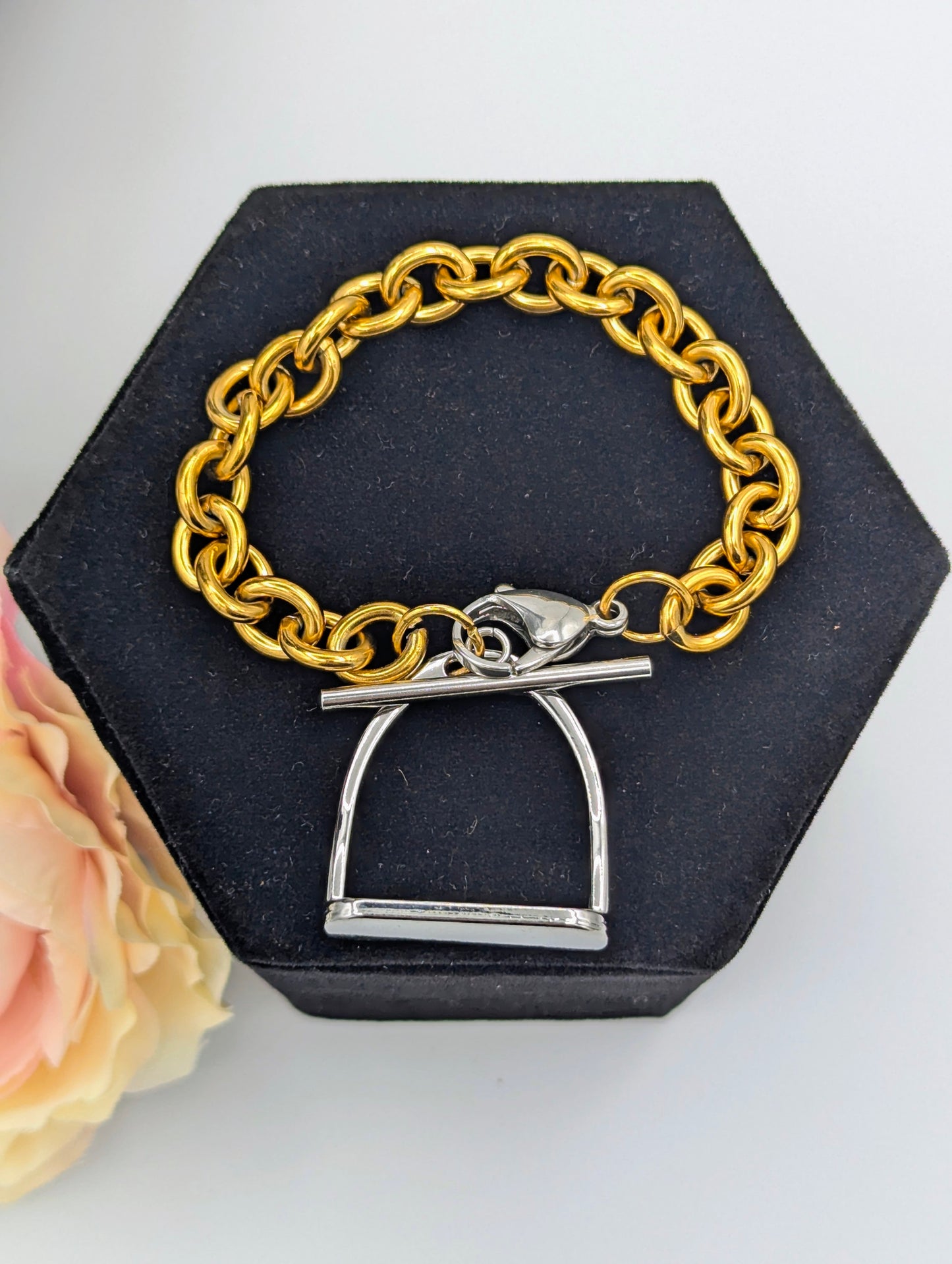Chunky two‑tone English stirrup bracelet with large gold‑tone chain links and a silver stirrup‑style clasp displayed on a black hexagonal jewelry stand, accented by a soft pink‑and‑cream flower.