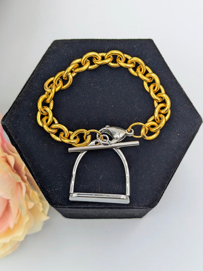 Chunky two‑tone English stirrup bracelet with large gold‑tone chain links and a silver stirrup‑style clasp displayed on a black hexagonal jewelry stand, accented by a soft pink‑and‑cream flower.