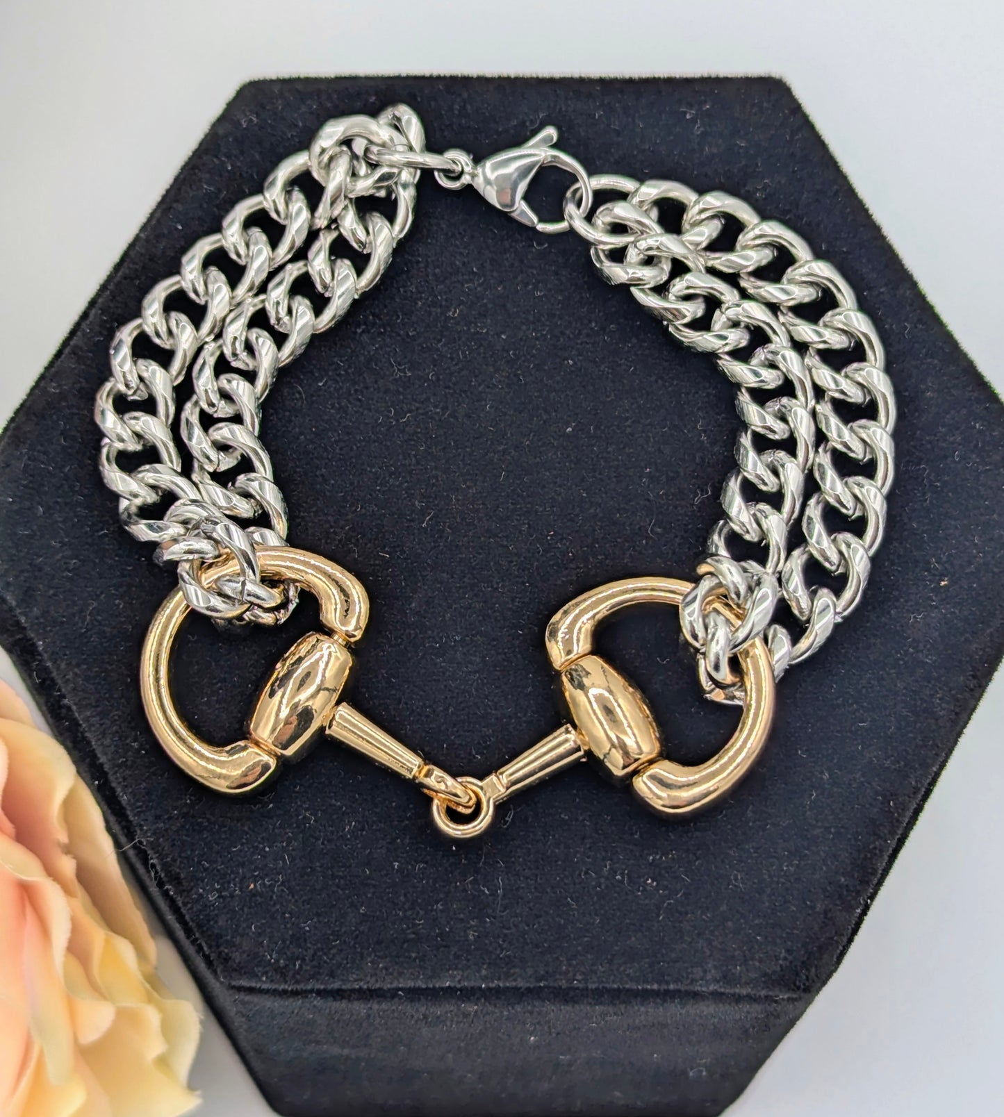 Two‑tone chunky snaffle bit bracelet with silver chain links and a gold‑tone snaffle bit centerpiece displayed on a black hexagonal jewelry stand, accented by a soft peach flower.