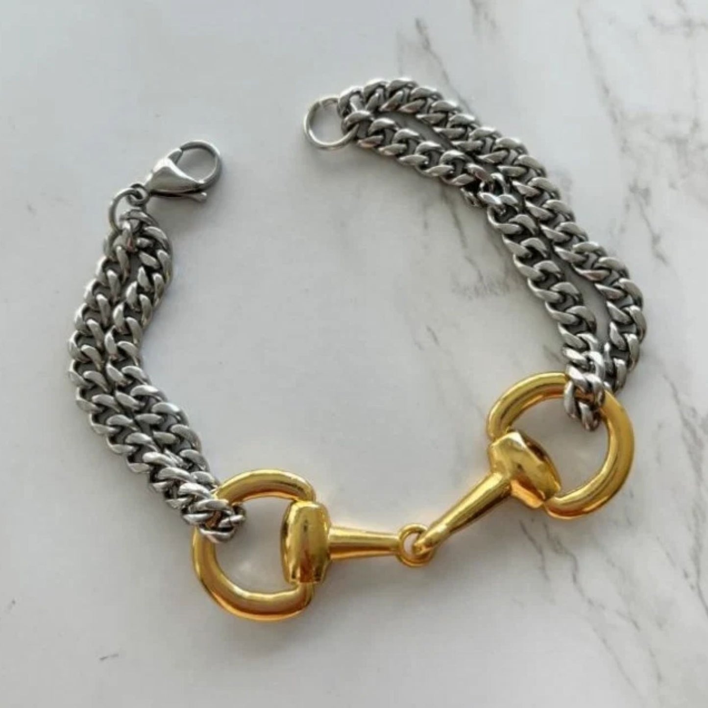 Two‑tone equestrian snaffle bit bracelet with a gold‑tone bit centerpiece and chunky silver stainless‑steel chain displayed on a light marble surface.