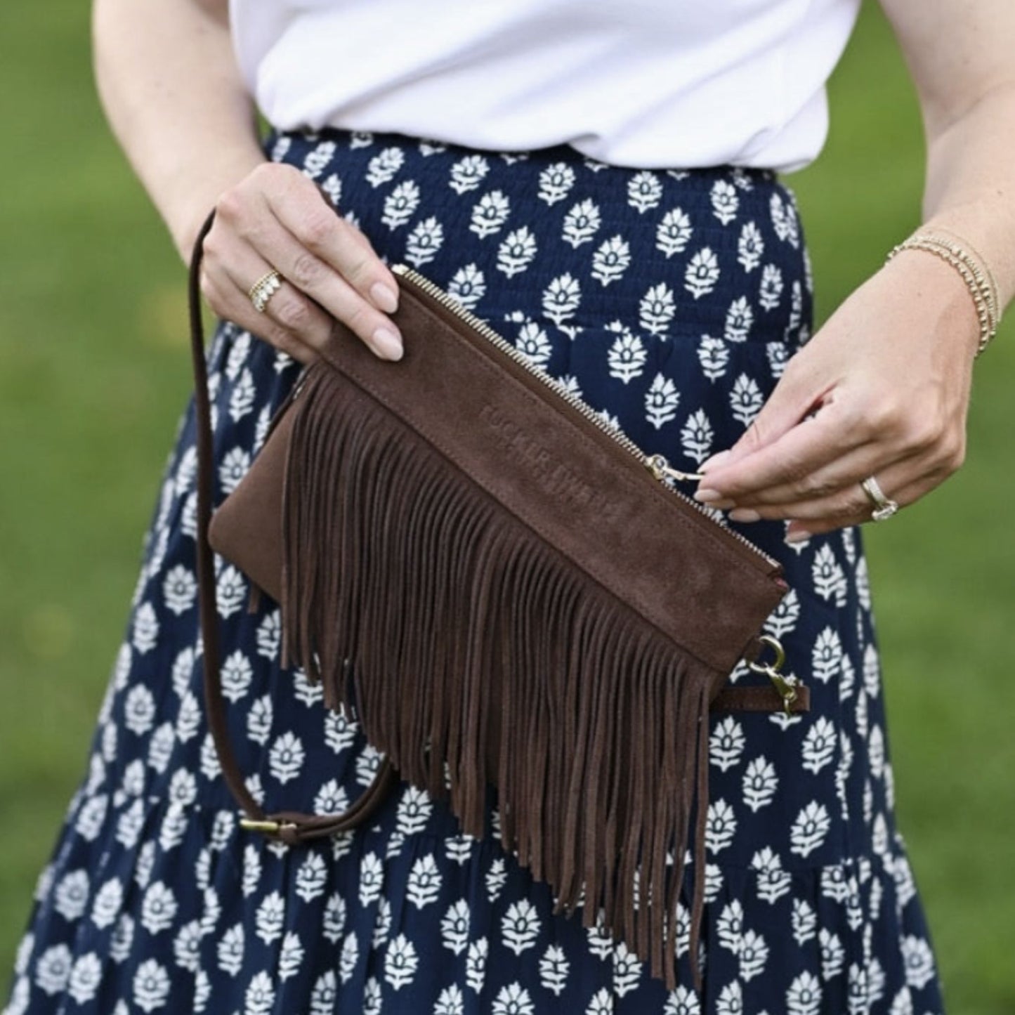 A person outdoors holding a brown suede fringe clutch bag with a white top and a navy blue floral skirt.